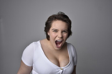Close up portrait of young plus sized woman with short brunette hair,  wearing a white shirt, with over the top emotional facial expressions against a light studio background.  