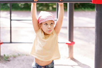 little girl exercising on a horizontal bar in a city park on a summer day