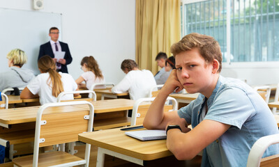Sad upset teenage schoolboy sitting separately in classroom during lesson in college