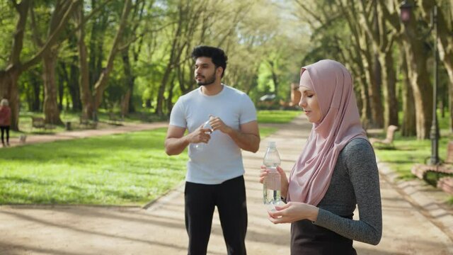 Side view of attractive woman in hijab drinking water while handsome arabian guy standing behind. Young couple in sportswear relaxing after workout on fresh air. Arabian family at park with water