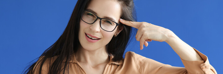 Young woman in glasses holding index finger near forehead on blue background
