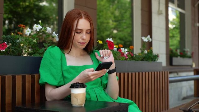 Attractive stylish young woman sending audio voice message on cellphone sitting at table with coffee cup in outdoor cafe terrace in sunny summer day. Happy redhead lady talking to mobile assistant.