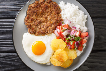 Authentic Silpancho Bolivian food from rice potatoes, beef, and fried eggs are topped with fresh salsa closeup in the plate on the table. Horizontal top view from above
