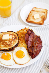 Full American Breakfast with Bacon, Hash Browns, Eggs and Pancakes on a plate on a white wooden table, side view.