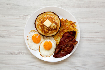 Full American Breakfast with Bacon, Hash Browns, Eggs and Pancakes on a plate on a white wooden background, top view.