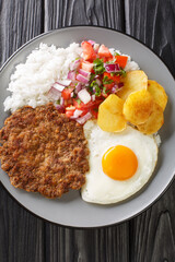 Silpancho is an incredible Bolivian meal made of rice and is topped with a pan fried burger, salsa, fried potatoes, and egg closeup in the plate on the table. Vertical top view from above