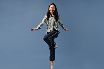 Happy brunette girl in black and white striped shirt and wide trousers smiling and jumping on blue isolated background..