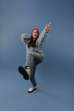 Positive long dark haired lady with red hat and glasses in trendy sweater, blue jeans and light cool boots raising her leg and arm..