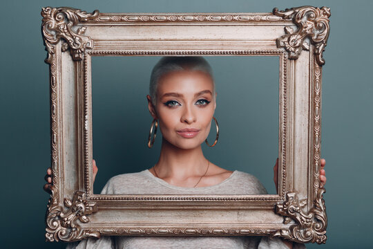 Millenial young woman with short blonde hair holds gilded picture frame in hands. Female face portrait