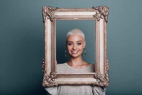 Millenial young woman smiling portrait with short blonde hair holds gilded picture frame in hands front of her face