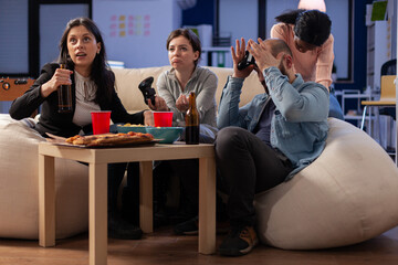 Team of multi ethnic friends losing tv game on console after work at office. Cheerful diverse group play with joystick while eating snacks pizza chips and drinking beer alcohol from cups