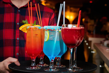The waiter holds a tray with Multicolored alcoholic and non-alcoholic cocktails with ice and straws