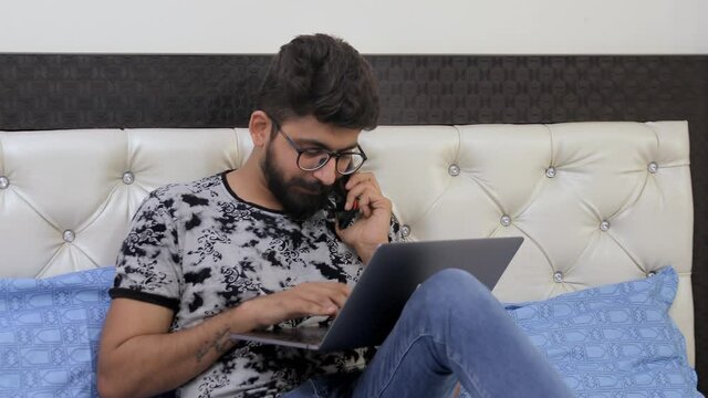 Young man busy over a phone call while doing work from home on his laptop. Bearded Indian guy in glass spectacles  working on his PC while talking on a smartphone - technology concept