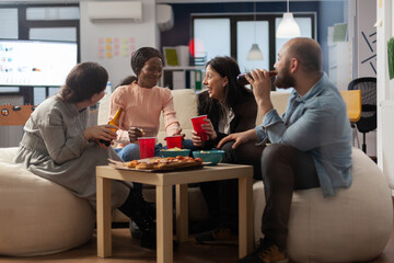 Multi ethnic colleagues having fun after work at office while eating pizza chips and drinking beer from cups and bottles. Diverse friends enjoying celebration party with snacks on table