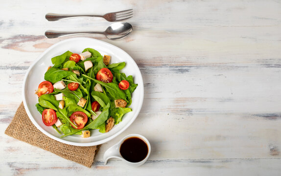 Fresh spinach salad with tomato, small pieces of herb roasted chicken in white ceramic dish on brown sack cloth, together with balsamic vinegar and metal utensil on white wooded table