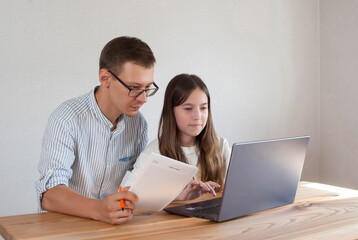 Young dad helps little daughter schoolgirl on distance learning sitting at table with computer at home