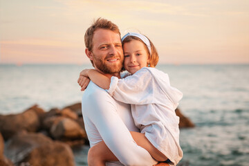 Father's Day. Portrait of a smiling father hugging his preschool-age daughter in his arms. In the background, the sea and the sky. The concept of happy fatherhood and adoption of children - obrazy, fototapety, plakaty