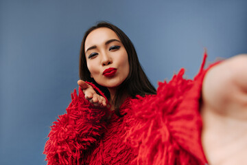 Bright gorgeous asian lady with dark hair and beautiful makeup in fluffy red sweater blowing kiss and taking selfie on blue backdrop..