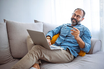 Man in living room with laptop and credit card smiling. Smiling man holding a credit card and using laptop at home. On-line shopping. Photo of mature man a application for payment from home