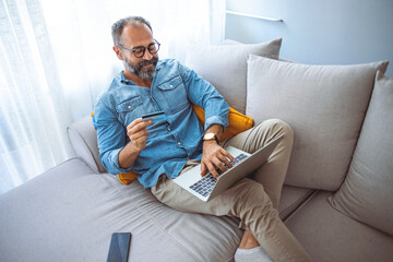 Smiling mature man looking on credit card and providing online payment while sitting on couch at home. Online shopping concept. Man at home buying on internet