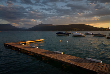 Small wooden dock and motorboats on Annecy Lake water with a sky full of clouds at sunset with...