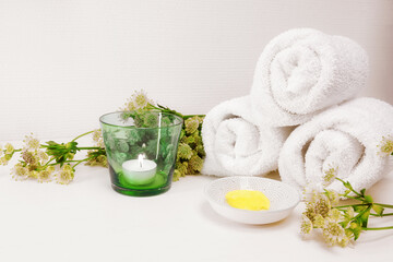 Fototapeta na wymiar Cosmetic moisturizer or emollient for pedicure and soft foot skin, white towels, candle and some green flowers on a light background with large copy space