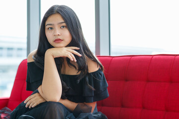 Front view portrait shot of a beautiful smiling young junior teenager in a black dress sitting on the couch and looking at the camera in the studio. Beautiful adolescent girl in a prom party