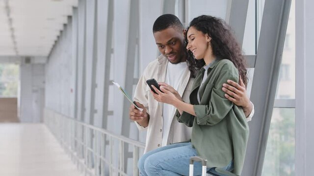 Multiracial couple african man and hispanic woman with plane tickets passports sitting in airport terminal using navigator app mobile phone watching photo from trip browsing network with smartphone