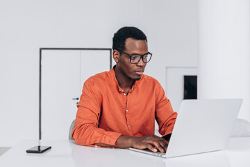 Concentrated young African-American guy in orange clothes and glasses works on modern laptop at comfortable workplace closeup