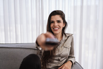 Fototapeta na wymiar A positive smiling optimistic young Latin woman sit indoors at home watch tv holding remote control on sofa.