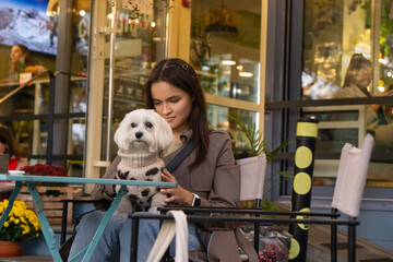 A young brunette girl in a trench coat with a dog dressed in a sweater is sitting in a cafe in an...
