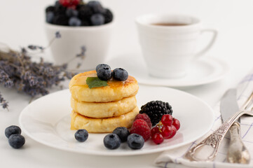 Blurred image of cheesecakes with honey and fruits on a white plate, in the background a cup of coffee, a plate with fruits and a bouquet of lavender.Proper nutrition.
