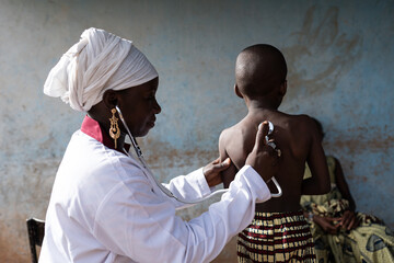 Young smiling doctor in a clean white uniform auscultating a little boy's lungs sounds with a modern stethoscope on his bare back
