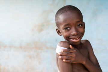 Portrait of a smiling and confident looking bare chested skinny little African boy with a large patch on his shoulder after being vaccinated against childhood infectious disease - obrazy, fototapety, plakaty