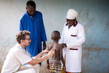 White medical volunteer auscultating the lungs of a skinny little African boy under the supervision...