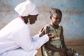 Black nurse gently placing a digital thermometer under the arm of a smiling little African girl...