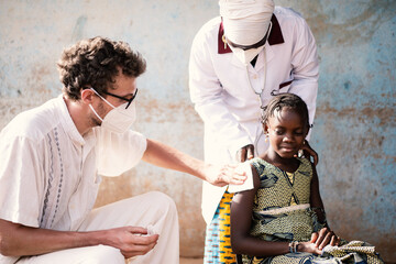 Caucasian physician with protective face mask disinfecting the injection site of a doubtful and tense looking little African girl supported by a local nurse