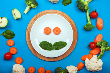 Yummy toddler food in white plate on blue background. Concept first complementary food made from fruits and vegetables. Smilie made from carrots and spinach.