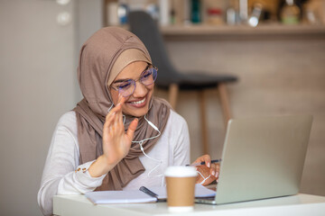 Smiling Muslim girl student wear wireless headphone study online with skype teacher, happy young...