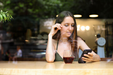 a young girl in summer clothes uses the phone while sitting in a cafe and drinking coffee or tea. woman texting on social networks, view from the street through glass