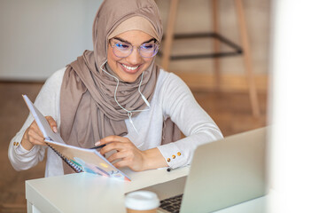 Muslim woman in wireless headphones sit at desk at home working on modern laptop, young ethnic...