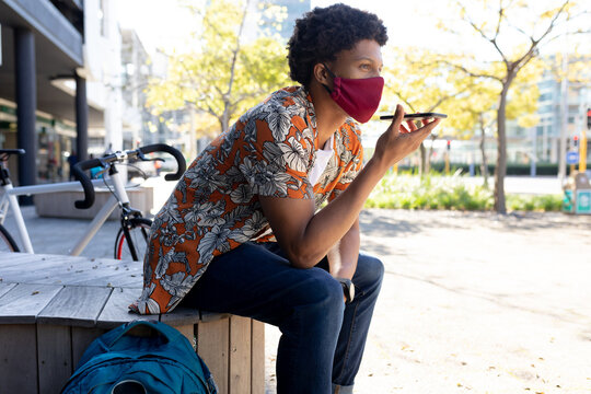 African american man in city wearing face sitting and using smartphone