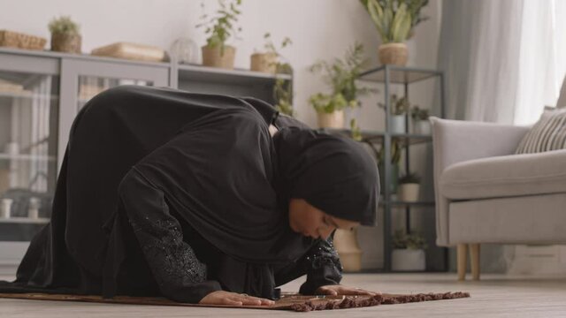Slowmo shot of concentrated young Muslim woman praying indoors at home sitting on carpet