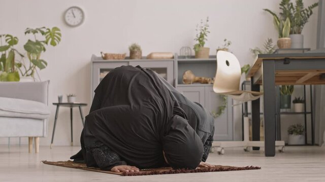 Slowmo shot of young Muslim woman in traditional black dress and hijab praying on carpet at home