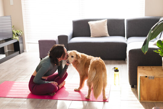 Caucasian woman practicing yoga, sitting on yoga mat kissing her dog