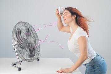 A red-haired Caucasian woman chills by the electric fan and drinks a cold drink. Climate control in...