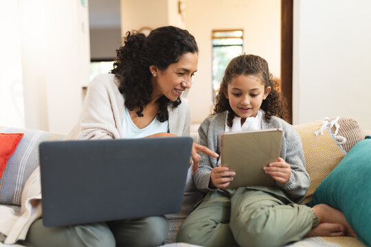 Smiling mixed race mother and daughter sitting on sofa, using laptop and tablet