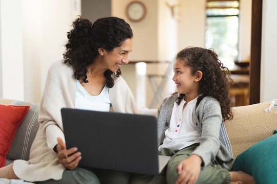 Smiling mixed race mother and daughter sitting on sofa and using laptop