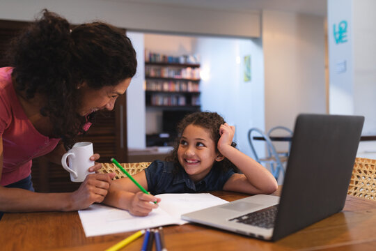 Smiling mixed race mother holding coffee, helping her daughter do homework