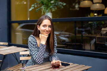 caucasian girl looking at the camera and smiling while sitting at the table of modern outdoor cafe on the background of the window